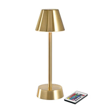 The wireless LED lamp ZELDA in brass by Duni GmbH with a conical shade and cylindrical rod stands next to a small remote control with colorful buttons and, together with a handmade vase, adds elegance to the room.