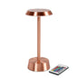 The NOUR wireless LED lamp in rose gold from Duni GmbH has a round base and flat top for a modern style. Next to the lamp is a remote control with colorful buttons.