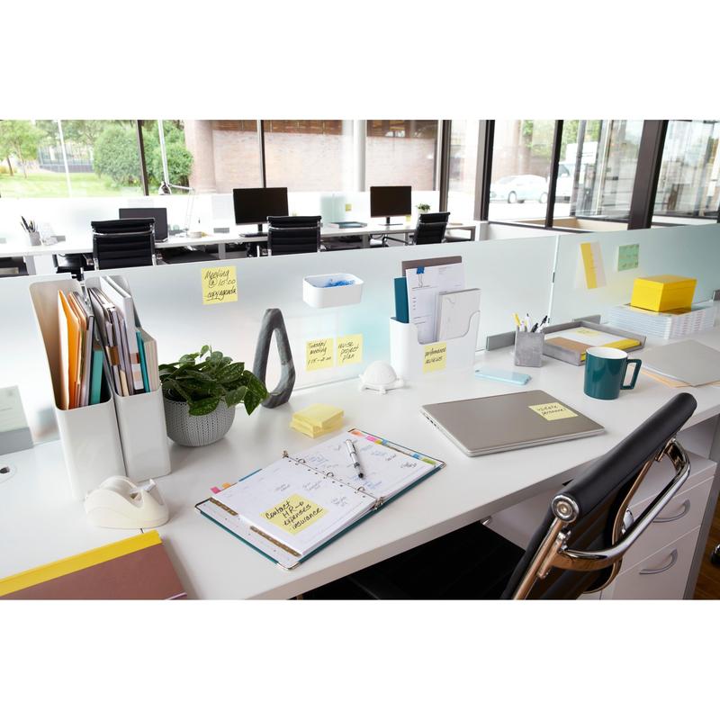 A modern office desk with a laptop, planner, coffee mug, plant, stationery, and 3M Post-it® Notes yellow (76x76 mm, 100 sheets/block), ideal for quick notes. The files are organized and large windows offer a view outside; the desks are arranged in a row.