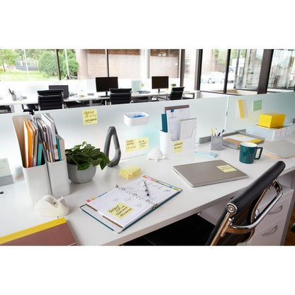 A modern office workspace with a desk with folders, laptop, planner, and 3M Deutschland GmbH Post-it® Notes yellow (76 mm x 127 mm, 100 sheets/block, 12 blocks/pack), with large windows and empty chairs in the background.