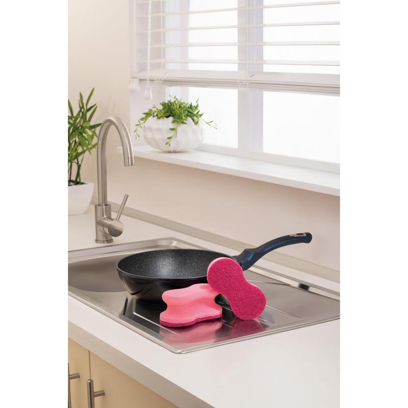 A black frying pan lies in a kitchen sink, on the edge of which are two pink Scotch-Brite® Delicate Color Comfort non-scratch cleaning sponges (2 pieces, 3M Deutschland GmbH); the white countertop and potted plants nearby complete the scene.