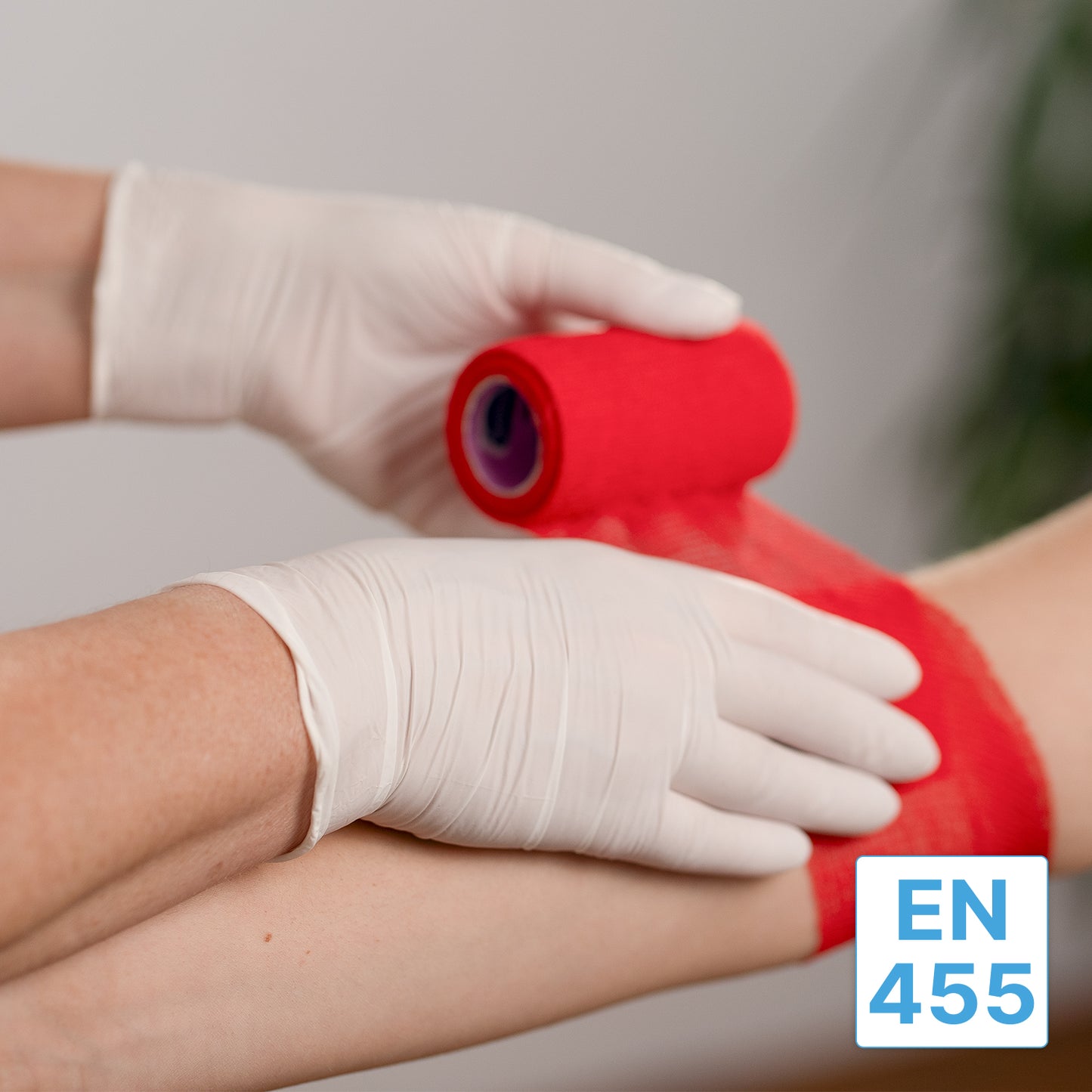 A person wearing ARNOMED NITRIL WHITE 200 nitrile gloves from ARNOWA GmbH wraps a red bandage around another person's arm. In the lower right corner, a blue and white "EN 455" label is visible.