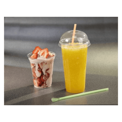 A Starpak 50 drinking cup "Hurricane" PET crystal-clear is filled with orange slush and stands next to a plastic cup with strawberries in yogurt. A green straw lies in front of them on the gray surface.