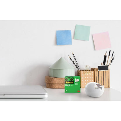 A desk with a closed laptop, a 3M Deutschland GmbH Scotch™ Karim Rashid tape dispenser and a box of tape, containers for pens and pencils, a small house-shaped ornament, and three colorful sticky notes on the white wall.