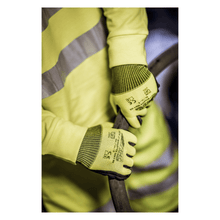 A person wearing a yellow safety vest and NITRAS NYLOTEX SOFT HV nylon gloves from AS Arbeitsschutz GmbH, neon yellow, partially coated – with black text and safety symbols – is holding a thick, dark cable or hose.