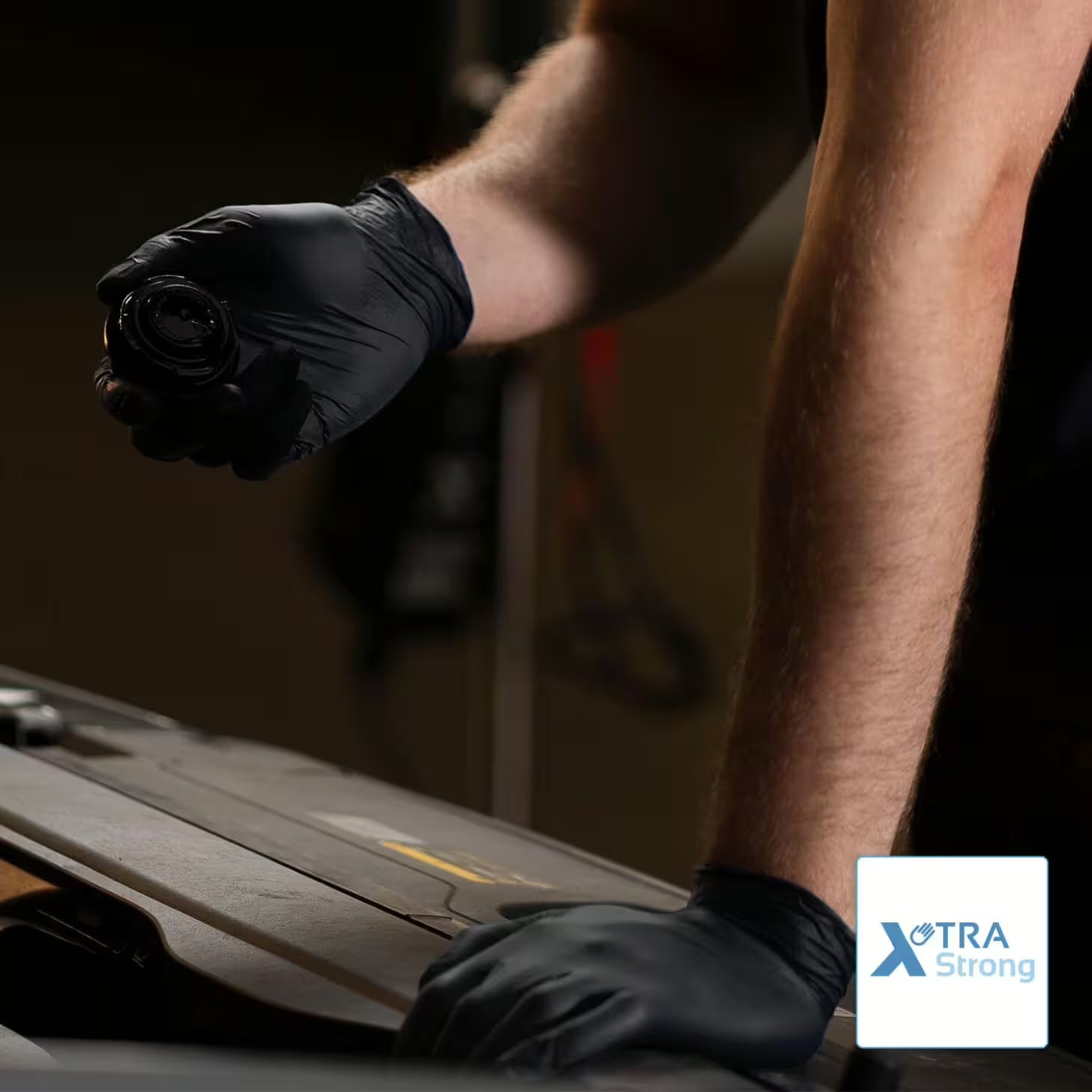 A person wearing ARNOMED NITRIL XTRA PURE BLACK disposable gloves from ARNOWA GmbH holds a round car part over an open engine, with the arm resting on it. In one corner is the "XTRA Strong" logo.