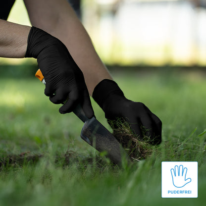 A person wearing ARNOMED NITRIL BLACK XTRA STRONG extra thick disposable gloves made of nitrile in black from ARNOWA GmbH uses a small trowel to dig outdoors. In the corner, a blue "Powder-free" hand symbol is visible.