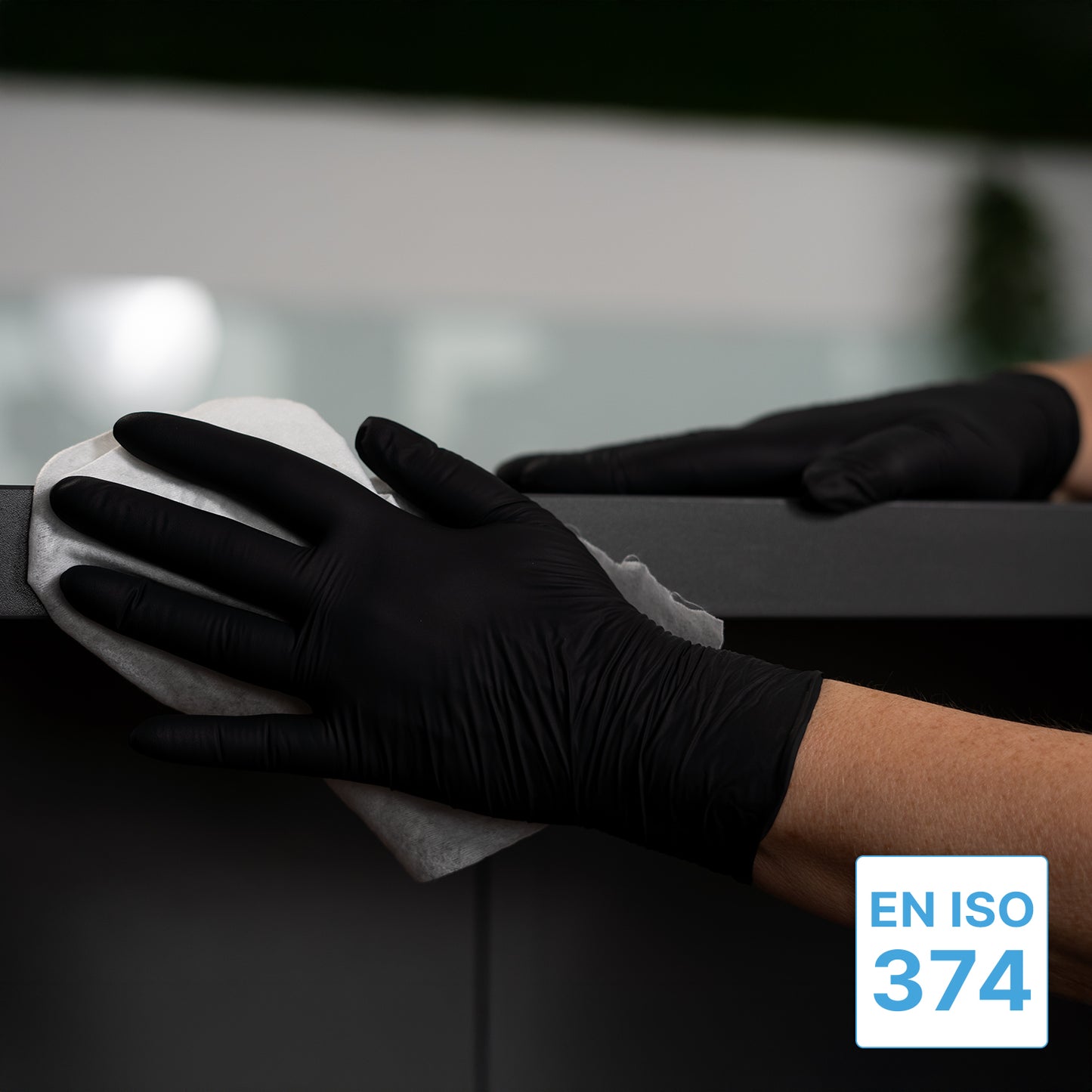 A person wearing ARNOMED NITRIL BLACK XTRA STRONG extra thick black nitrile disposable gloves from ARNOWA GmbH wipes a dark surface with a white cloth. In the lower right corner, the blue-white label "EN ISO 374" appears.