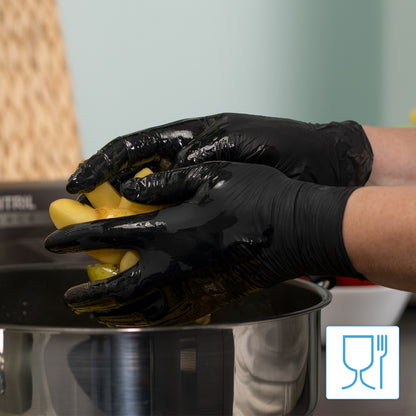 A person wearing ARNOMED NITRIL BLACK 200-pack black nitrile gloves from ARNOWA GmbH holds yellow peppers over a stainless steel pot. The latex- and powder-free gloves ensure safe handling of food; utensil and glass symbols appear in the corner of the image.
