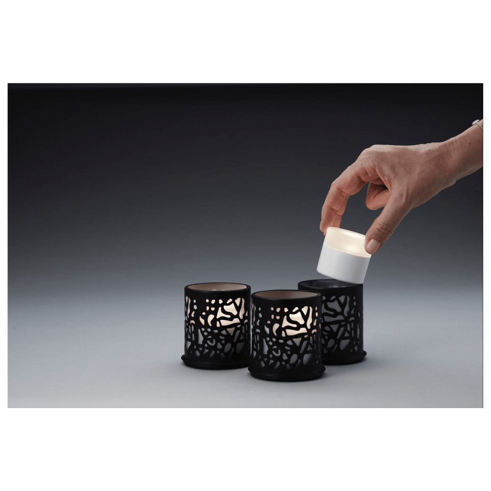 A hand places a small cylindrical candle into one of three Twine 75 x Ø 75 mm Black candle holders from Duni GmbH, which are adorned with elaborate cut-outs and are ideal for modern design. The table decoration is placed against a gradient background from dark to light gray.