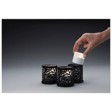 A hand places a small cylindrical candle into one of three Twine 75 x Ø 75 mm Black candle holders from Duni GmbH, which are adorned with elaborate cut-outs and are ideal for modern design. The table decoration is placed against a gradient background from dark to light gray.