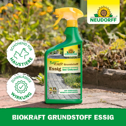 A spray bottle with Neudorff BioKraft Base Material Vinegar from the Neudorff Shop stands on a garden path, surrounded by grass and flowers. The label emphasizes pet safety and effective weed removal for clean terraces.