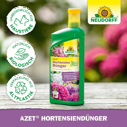 A green bottle of Neudorff Shop Azet Hydrangea Fertilizer liquid with floral images on the label, depicted with symbols for pet-safe, organic fertilizer, and bioplastic. The background shows blurred flowers.