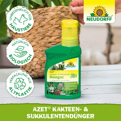 A green plastic bottle of Neudorff Shop Azet Cactus & Succulent Fertilizer liquid stands on a wooden table in front of a potted cactus. Three symbols indicate that it is pet-friendly, organic, and made from recycled plastic.