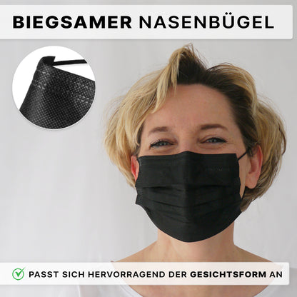 A person with short blonde hair wears an ARNOMED MOUTH PROTECTION TYPE IIR surgical mask with rubber bands from ARNOWA GmbH. The black disposable mask with a flexible nose bridge is seen in a close-up, with German text indicating the adjustable fit.
