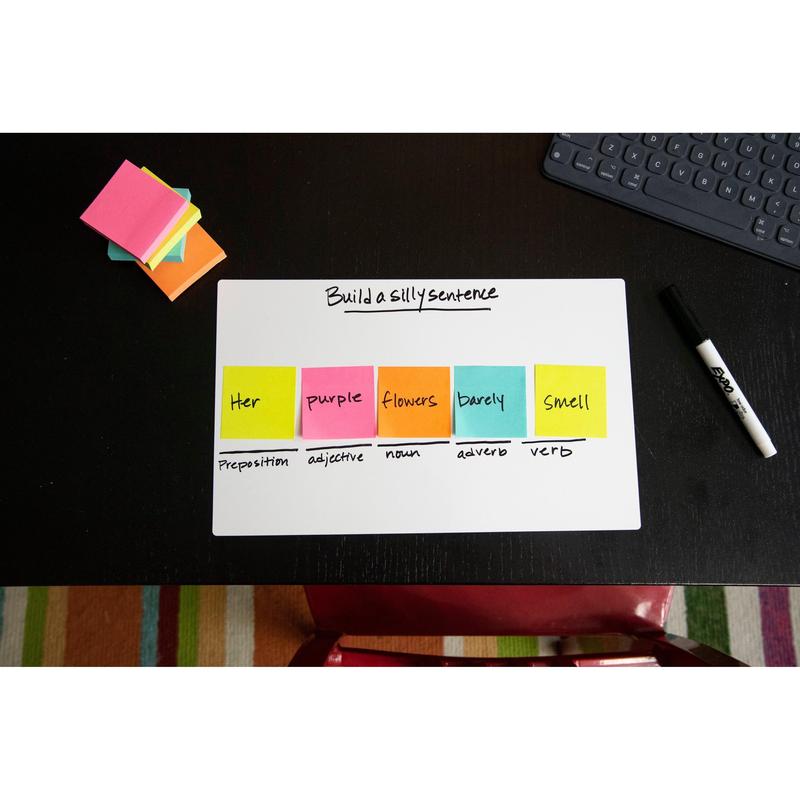 On a black desk stands a whiteboard with the inscription "Build a silly sentence". The Post-it® Notes Cube, Ultra Colors, 76x76 mm, by 3M Deutschland GmbH, display the parts of speech. Also on the desk: a dry-erase marker, a notepad, and a keyboard.