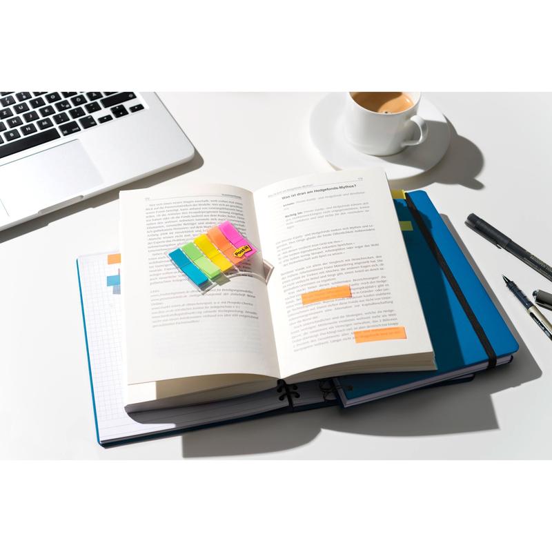 On a well-lit desk lies an open book labeled with 3M Deutschland GmbH Post-it® Index Tabs Mini (11.9 x 43.2 mm, 100/dispenser), over a blue notebook and laptop, with pens and coffee on the white surface nearby.