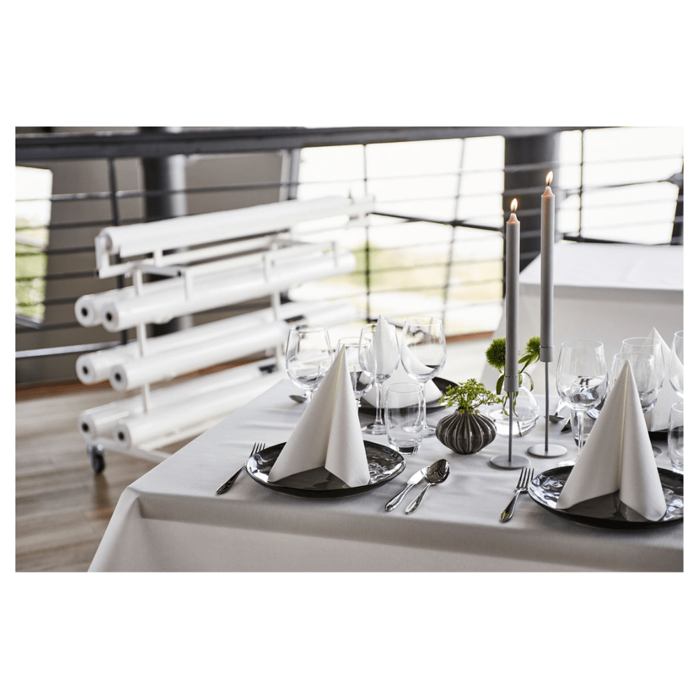 On a beautifully set dining table stands a fresh, white Duni Dunicel® tablecloth roll from Duni GmbH, offering a fabric-like texture. Triangular folded napkins on dark plates accompany wine glasses. Tall candles and small floral arrangements serve as table decorations, with rolled canvases visible in the background.