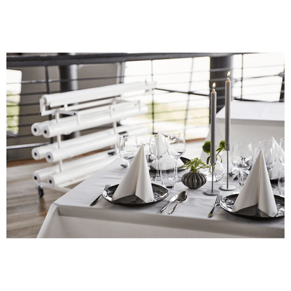 On a beautifully set dining table stands a fresh, white Duni Dunicel® tablecloth roll from Duni GmbH, offering a fabric-like texture. Triangular folded napkins on dark plates accompany wine glasses. Tall candles and small floral arrangements serve as table decorations, with rolled canvases visible in the background.