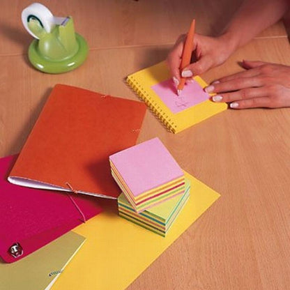 At a wooden desk, someone writes with an orange pen on a pink Post-it® Notes cube (76 mm x 76 mm, 450 sheets/block, 100% PEFC) from 3M Deutschland GmbH. Nearby are a red notebook, a green tape dispenser, a silver bell, and more colorful notes.