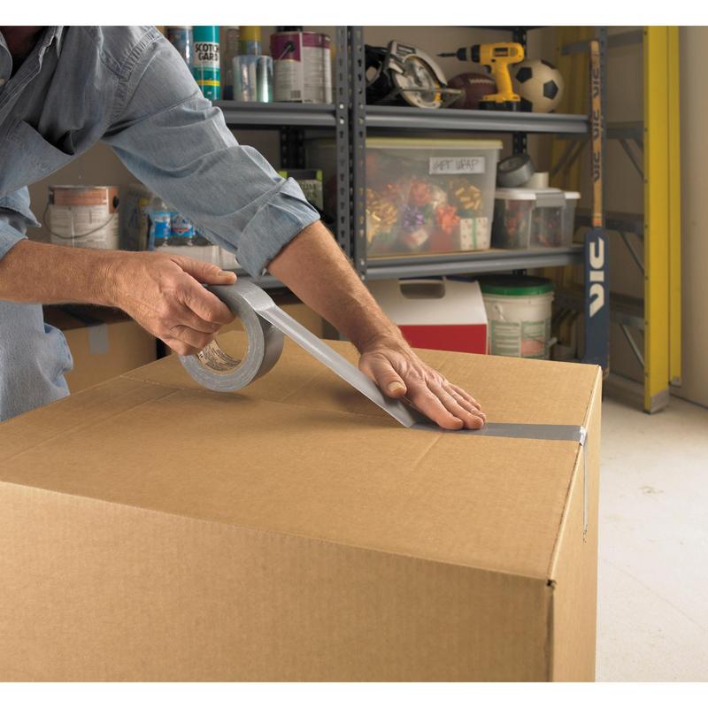A person seals a large box in a garage with Scotch® Universal Tape 2904 (50 m x 48 mm) from 3M Deutschland GmbH, with shelves of tools and accessories visible in the background.