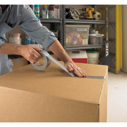 A person seals a large box in a garage with Scotch® Universal Tape 2904 (50 m x 48 mm) from 3M Deutschland GmbH, with shelves of tools and accessories visible in the background.