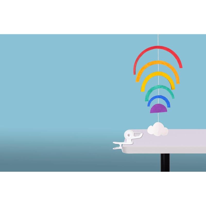 A colorful paper mobile with five rainbows and a white cloud hangs on the edge of a white table, secured by the 3M Deutschland GmbH Scotch™ Clip & Twist Tape Dispenser C19 with Magic™ Tape, against a light blue background.