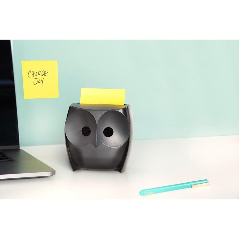 A black Post-it® Z-Notes Dispenser "Owl" (3M Deutschland GmbH) with yellow Super Sticky Z-Notes stands on a white desk, next to a laptop and a blue pen. A "CHOOSE JOY" note hangs on the wall against a light blue background.