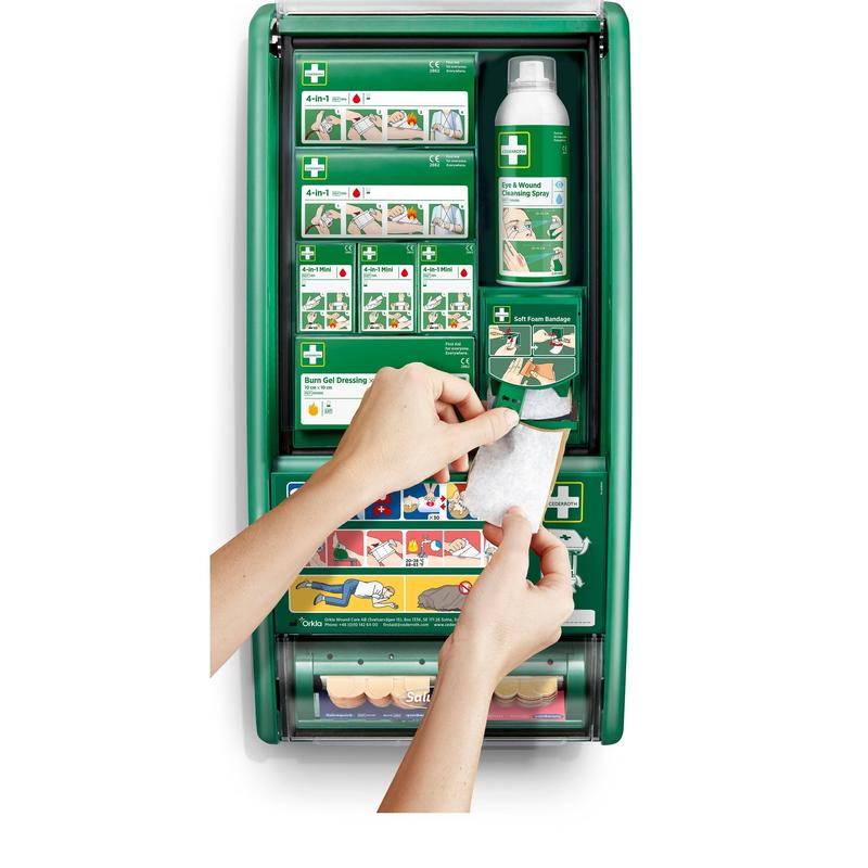 A person holds a bandage in front of a Cederroth First Aid Station by Orkla Wound Care AB, which is stocked with bandages, burn gel, saline solution, and bindings. Illustrated instructions can be seen on the compartments inside.