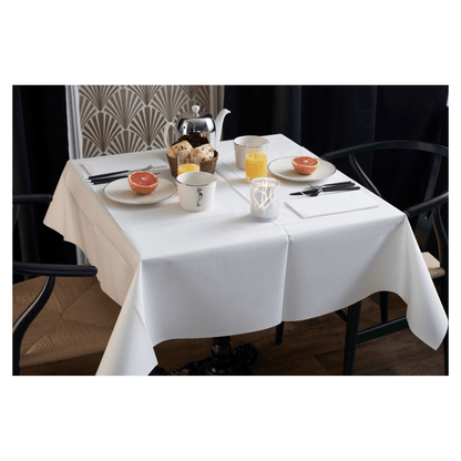 The breakfast table is elegantly set with a white tablecloth, two chairs, and a vase with a geometric pattern. On the table are grapefruit halves, pastries, tea, and orange juice. A Duni Dunilin® napkin (48x48 cm) from Duni GmbH adds charm and functionality to the atmospheric morning setting.