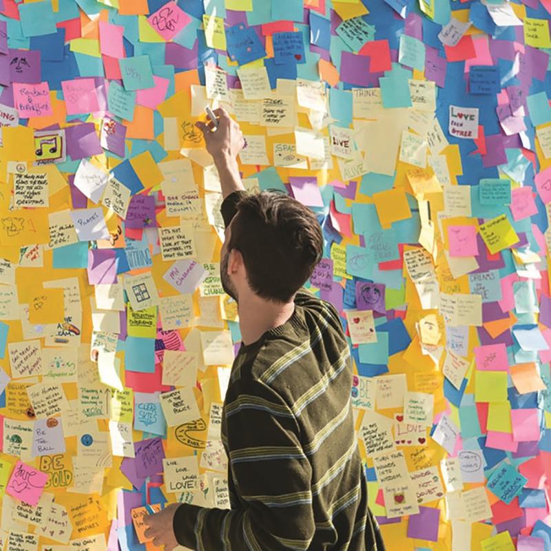 A person in a striped shirt stands by a wall with handwritten messages and drawings and reaches for a 3M Deutschland GmbH Post-it® Notes Cube in Ultra Colors (76x76 mm, 450 sheets, 100% PEFC) to place it among the colorful notes.