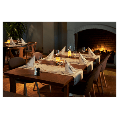 A cozy dining area features two wooden tables with wine glasses and Duni Bio Dunisoft® napkins next to flickering candles. Dark chairs surround the tables, enhanced by a burning fireplace. Plush seating and a plant contribute to the nature-inspired design.