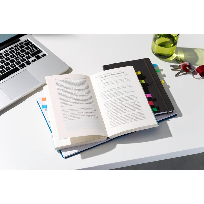 An open book with 3M Post-it® Index Flags, Mini, in various colors and blue/yellow index arrows lies on a notebook with more flags next to a laptop, a green water bottle, and keys on a white desk.