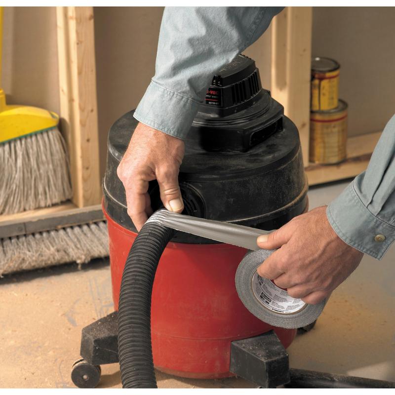 A person wraps Scotch® Universal Tape 2904 (50 m x 48 mm) from 3M Deutschland GmbH around a vacuum cleaner hose attached to a red shop vacuum. In the background, a broom and paint cans are visible.