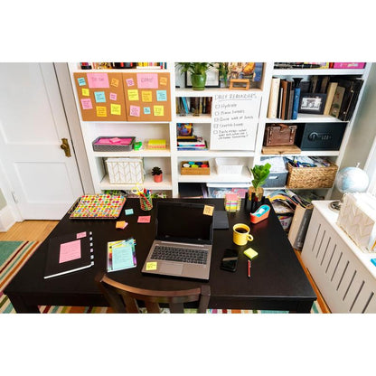 A desk with a laptop, notebooks, a phone, pens, a mouse, a yellow mug, and Post-it® Super Sticky Z-Notes (Boost Collection) from 3M Deutschland GmbH. Behind it are shelves with books, storage boxes, and a whiteboard with reminders and colorful sticky notes.