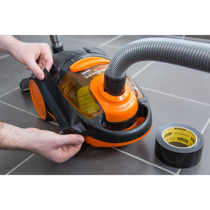 A person applies Scotch® Universal Tape 2904 from 3M Deutschland GmbH to the damaged side of an orange-black vacuum cleaner on a gray tiled floor, while a roll of tape lies nearby.
