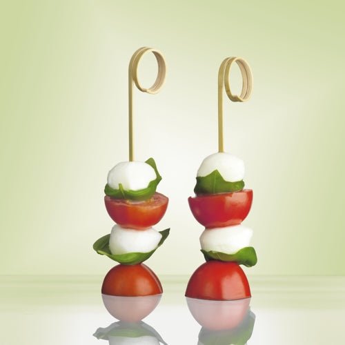 Two PAPSTAR 100 finger food picks "pure" from PAPSTAR GmbH made of high-quality plastic hold mozzarella balls, basil, and cherry tomatoes, decoratively against a soft green – ideal for parties and events.
