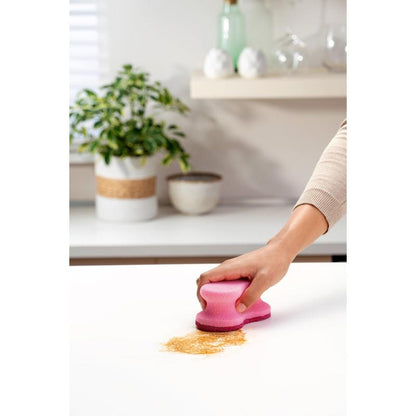 A hand uses a pink Scotch-Brite® Delicate Color Comfort non-scratch cleaning sponge (2 pieces, 3M Deutschland GmbH) to clean brown powder from a white countertop - perfect for delicate surfaces. Plant and bowls in the background.