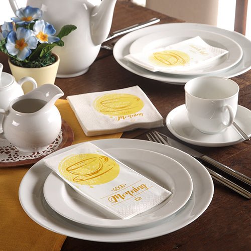 A neatly set table with white dishes, a teapot, blue flowers, and 3-ply PAPSTAR napkins (33 x 33 cm) by PAPSTAR GmbH with a yellow coffee cup and the inscription "Morning".