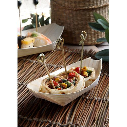 Three vegetable-filled flatbread rolls on skewers are served as finger food in a PAPSTAR 50 finger food wooden tray "boat" from PAPSTAR GmbH on a woven placemat. Another dish is blurred in the background.
