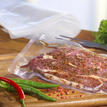 A marinated portion of meat lies in a transparent PAPSTAR 100 edge-sealed bag (PA/PE) from PAPSTAR GmbH on a wooden board with chili peppers and peppercorns, highlighting food storage. Leafy vegetables can be seen in the background.
