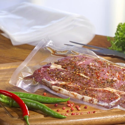 A raw marinated steak, sealed in a transparent Starpak 100 vacuum seal bag (PA/PE), lies on a wooden board next to red and green chili peppers, with tongs and leafy greens in the background.