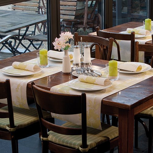 A wooden dining table for four with white plates, PAPSTAR 50 napkins "ROYAL Collection" 1/4-fold 40 x 40 cm green from PAPSTAR GmbH, green candles, glassware, salt and pepper shakers, and a vase with pink flowers by the window.