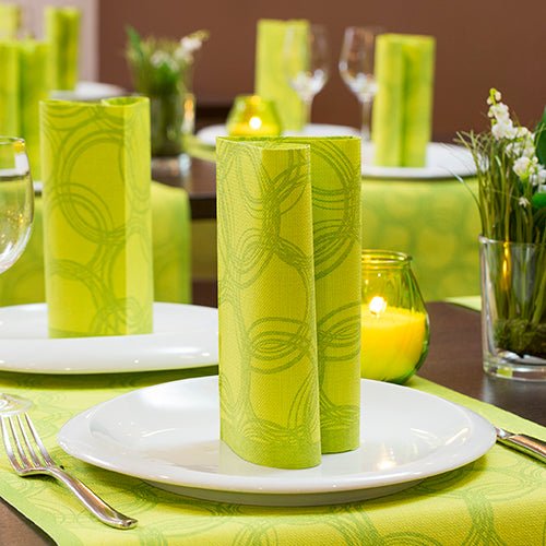 A dining table with white plates, PAPSTAR GmbH 50 napkins "ROYAL Collection" 1/4-fold 40x40 cm green upright, silver cutlery, wine glasses, flower vases, and green votive candles on a green runner for festive occasions.