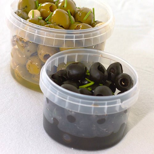 Two transparent Starpak 50 deli cups, round PP deli cups with original seal – one filled with green olives and herbs, the other with black olives and sprigs of herbs – stand on a light background.