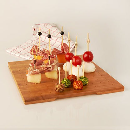 A wooden board with skewered snacks, served on the PAPSTAR holder for finger food picks, bamboo tray 25x30 cm from PAPSTAR GmbH. Cheese, sausage, tomatoes, and herb cheese balls are included; a checkered cloth can be seen in the background.