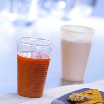 Next to a Starpak 25 drinking cup filled with orange juice (PP, 0.2 l, Ø 7.03 cm × 9.9 cm) stands a white plastic cup with a light drink, in front of it a part of a dinner plate can be seen - perfect for enjoying on the go.