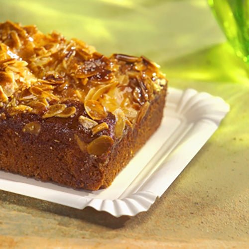 On the PAPSTAR cardboard plate rectangular 13x20 cm, white, "pure" for parties (12 pieces) by PAPSTAR GmbH rests a rectangular cake with shaved almonds - an eco-friendly, elegant serving option on a light surface with green glass behind.