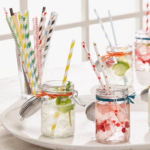Several mason jars with colorful drinks, ice, and PAPSTAR paper straws (Ø 6 mm) from PAPSTAR GmbH are arranged on a white tray. More patterned PAPSTAR paper straws fill the glasses behind on a white surface near a window.
