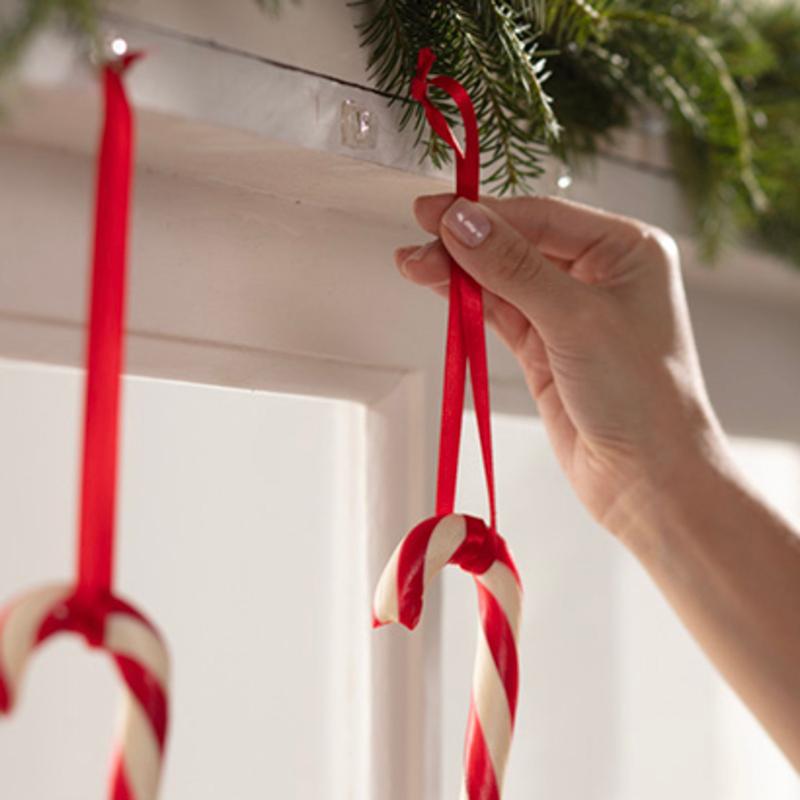 A hand hangs a red and white candy cane on a red ribbon on a white surface, with Command™ Mini Hooks, transparent, with transparent strips, 17006CLRFGN by 3M Deutschland GmbH. Another candy cane is already hanging next to it on green pine garland.